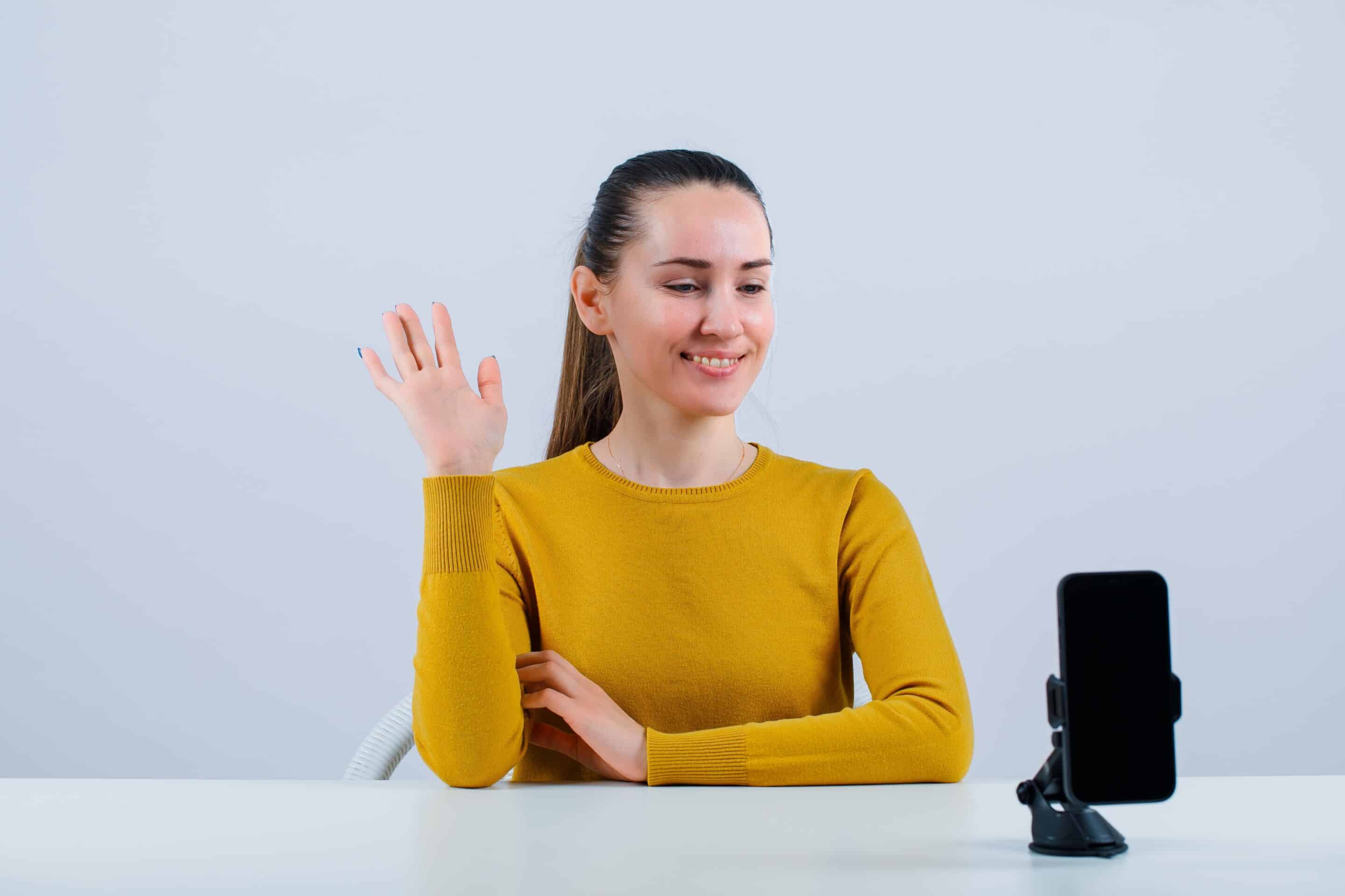 smiling-customer-girl-is-showing-hi-gesture-by-sitting-front-mobile-camera-white-background